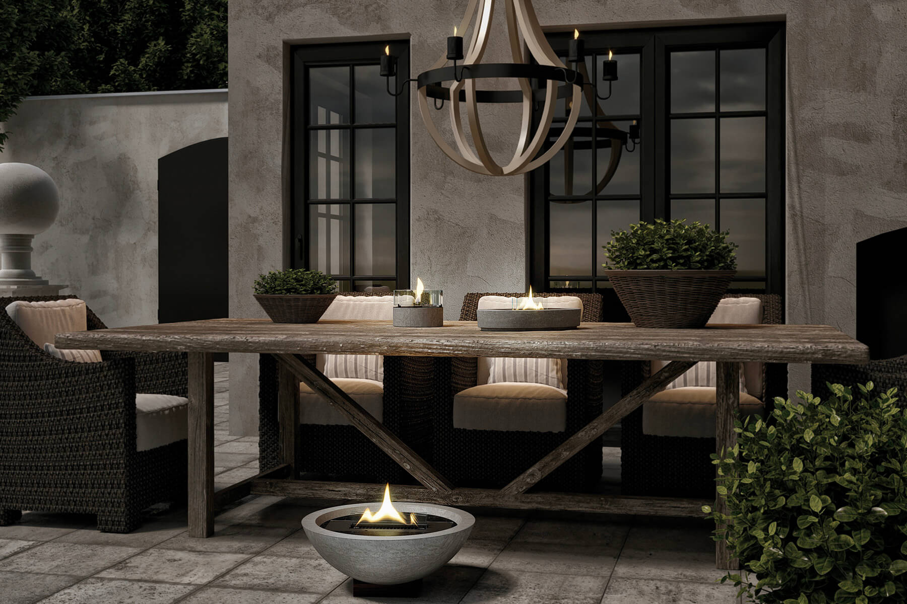 Terrasse am Abend: eine Feuerschale aus Beton am Boden und zwei Tischfeuer auf einer langen Holztafel unter einem Kerzenleuchter