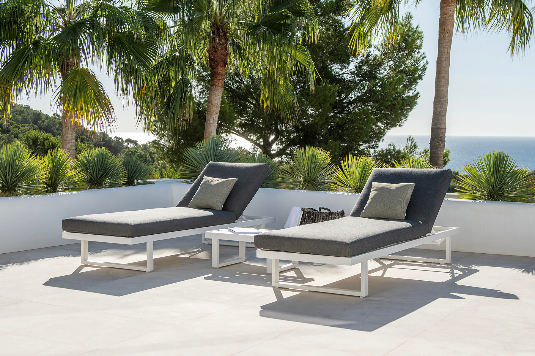 Zwei Sonnenliegen mit grauer Polsterung und wei&szlig;em Aluminiumgestell auf einer Terrasse, dahinter Palmen und das Meer