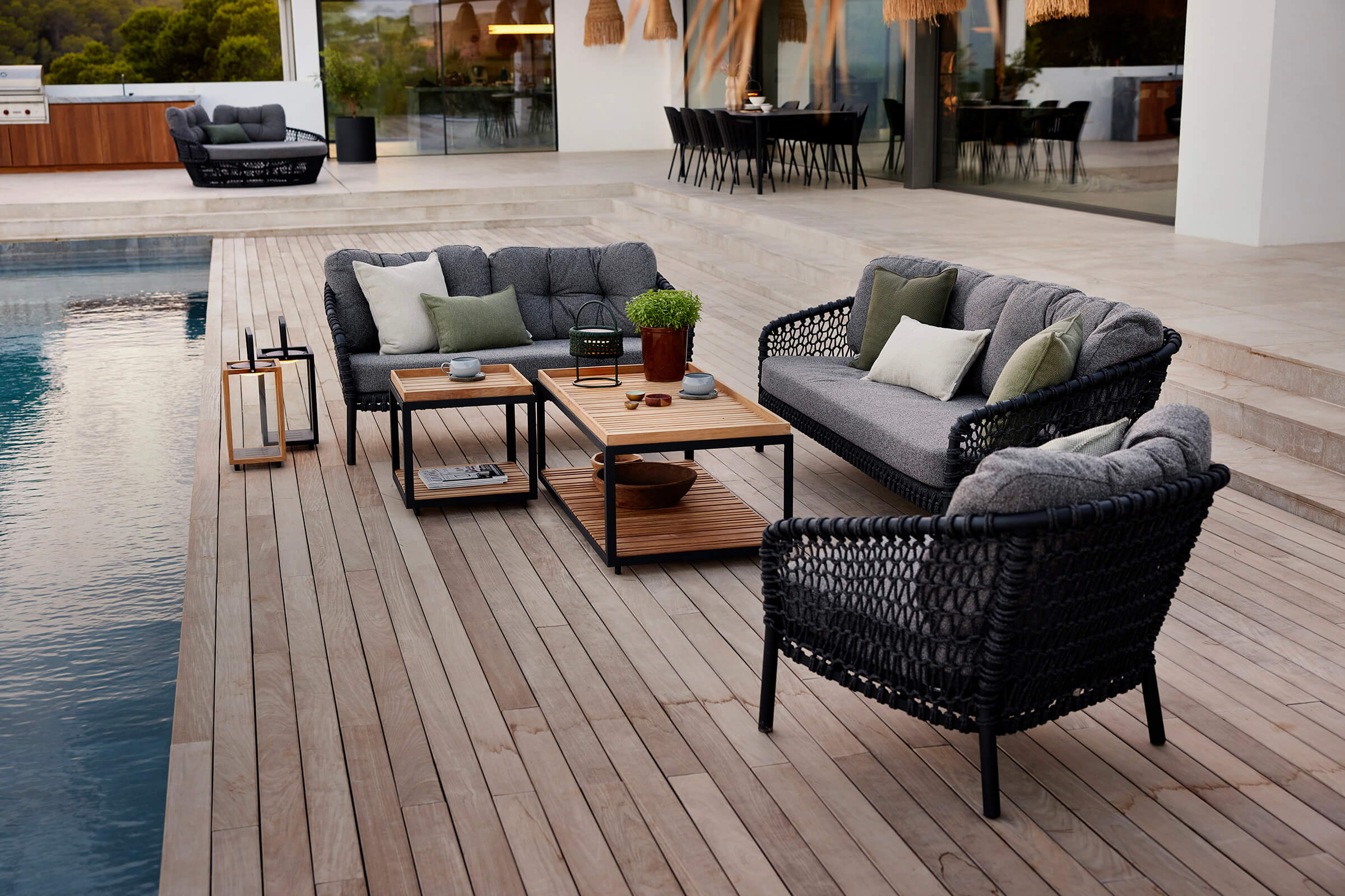 Lounge-Gruppe aus dunklem Seilgeflecht mit grauen Polstern auf einem Holzdeck am Pool, dahinter eine offene Terrasse mit Esstisch in der D&auml;mmerung