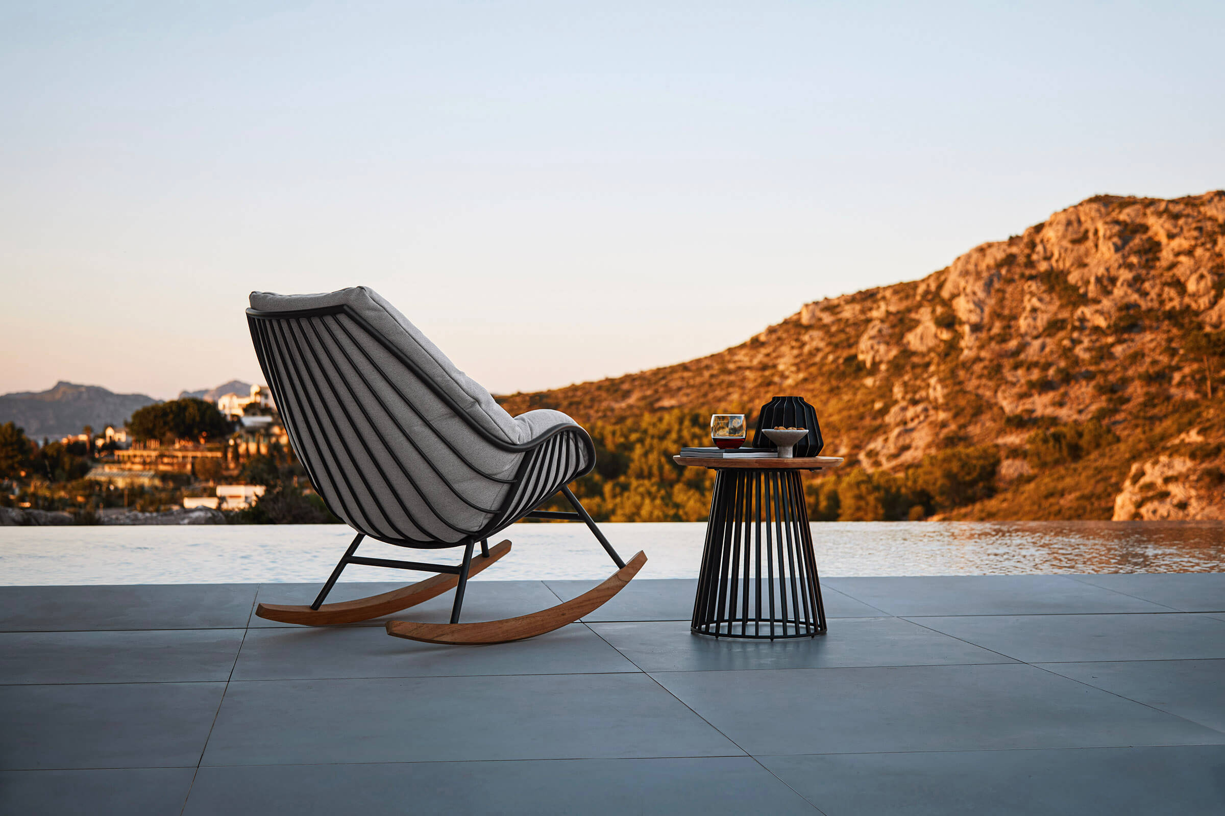 Schaukelsessel mit Kufen aus Teak und grauer Polsterung am Rand eines Infinity-Pools, dahinter eine H&uuml;gelkette im Abendlicht