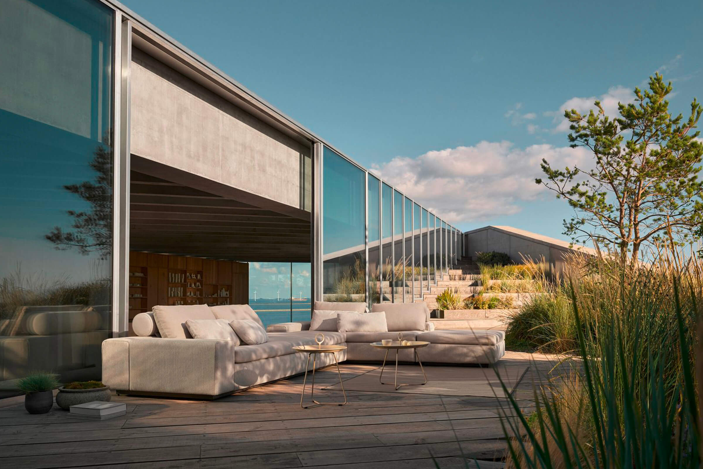 Helle Lounge-Landschaft auf einer Holzterrasse vor einem Haus mit raumhoher Verglasung, dahinter D&uuml;nengras und das Meer