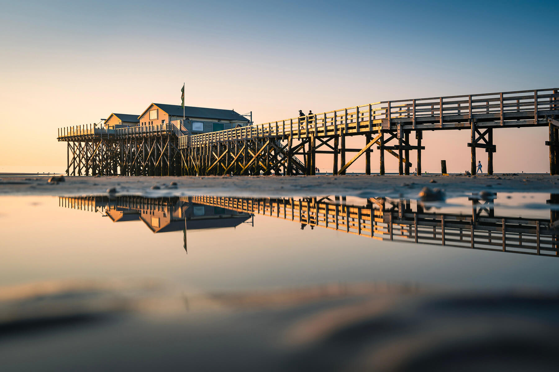 Pfahlbauten und Holzsteg am Strand von St. Peter-Ording im Abendlicht, gespiegelt in einem Priel