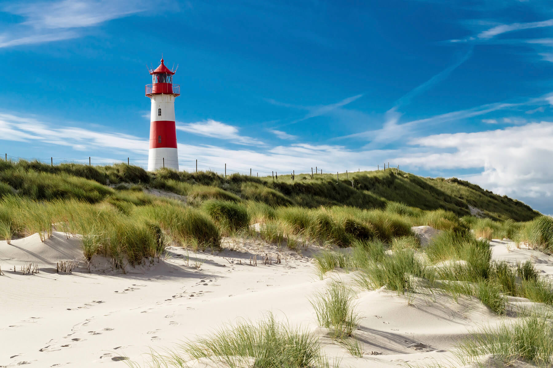 Rot-wei&szlig; geringelter Leuchtturm &uuml;ber einer D&uuml;ne mit Strandhafer, davor heller Sand unter blauem Himmel