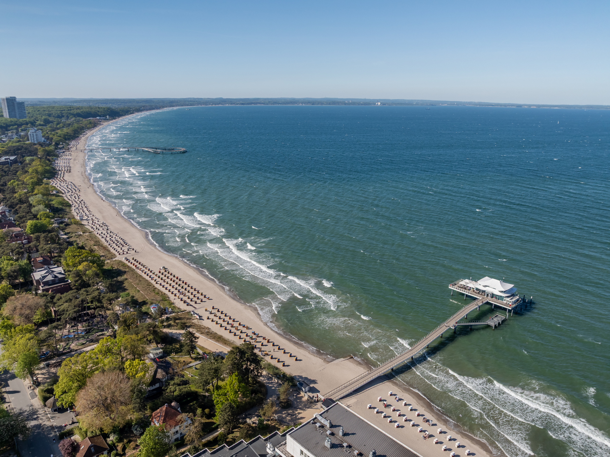Luftaufnahme der L&uuml;becker Bucht bei Timmendorfer Strand: die geschwungene K&uuml;stenlinie mit langer Reihe von Strandk&ouml;rben, dahinter Kiefern und Villen, rechts die Seebr&uuml;cke mit ihrem segelf&ouml;rmigen Dach